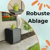 A garden storage box with a watering can on top, placed on a patio next to a wall with a window box of flowers. An inset image shows a close-up of the watering can on a shelf. Produktbild-Vorschau