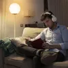 A man is sitting on a couch, reading a book and wearing headphones. A floor lamp is behind him. Podgląd zdjęcia produktu