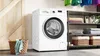 A white Bosch washing machine is shown in a laundry room setting, next to a wooden door and a shelving unit with folded towels and baskets. Produktbild-Vorschau