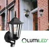 A black outdoor wall lantern with a motion sensor is shown mounted on a brick wall next to a white door. Another similar lantern is visible on the door. Produktbild-Vorschau