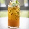 A close-up of a glass filled with a yellow-orange beverage, ice, passion fruit seeds, and mint leaves, garnished with a slice of passion fruit. Anteprima dell’immagine prodotto