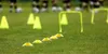 A sports field with training equipment, including cones and hurdles, with blurred figures of people in the background. Náhled obrázku produktu