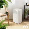 A white air dehumidifier is placed on a wooden floor next to a chair with a pillow and a scarf. Green plants are in the foreground and background. Voir les images du produit