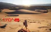 A person's legs are visible in the foreground, resting on sand dunes in a desert landscape. The sun is setting or rising, casting long shadows and illuminating the dunes. Produktbild-Vorschau
