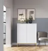 A white cabinet with gold legs is placed in a room with two framed pictures on the wall and a chair next to it. Produktbild-Vorschau