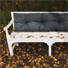 A white park bench with a gray cushion sits on the ground covered in fallen autumn leaves. Náhled obrázku produktu