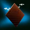 A square piece of dark chocolate with the Lindt logo embossed on it, floating against a dark blue and black gradient background. Small white pieces, possibly salt crystals, are scattered around the chocolate. Produktbild-Vorschau