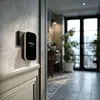 A wireless doorbell receiver is mounted on a wall in a hallway, with a doorway leading to a room with plants and a window. Náhľad obrázku produktu