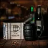 A black grenade-shaped mug and its packaging sit on a wooden table, with blurred bottles in the background. Voir les images du produit