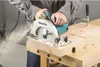 A person is using a Makita circular saw to cut a piece of wood on a workbench. Produktbild-Vorschau