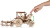 A wooden model tractor with a plow attachment is shown with a hand pressing down on the hood, indicating a feature or assembly step. Náhľad obrázku produktu