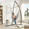 A woman is relaxing in a gray hammock chair indoors, listening to music on headphones and looking at her phone. The scene is set in a living room with a bookshelf, a side table with a drink, and a window with a view of greenery. Produktbild-Vorschau