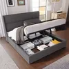 A grey upholstered bed with its storage compartment open, revealing various storage boxes and pillows inside. The bed is in a room with a window and furniture in the background. Produktbild-Vorschau