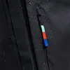 A close-up view of a black backpack with water droplets on the fabric, showing a zipper pull with aqua, red, and blue segments. Produktbild-Vorschau