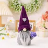 A decorative gnome figurine with a purple hat and a bouquet of purple flowers, placed on a table with other decorations. Produktbild-Vorschau