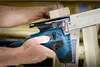 A person is using a blue and black Bosch jigsaw to cut a piece of wood. Voir les images du produit