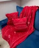 A red blanket with white patterns and two red pillows with white patterns are placed on a blue sofa. Produktbild-Vorschau