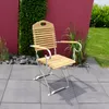 A wooden and metal garden chair is placed on a tiled patio in front of a red wall and green foliage. Produktbild-Vorschau