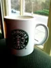 A Starbucks mug sits on a dark green surface, with a blurred window view in the background. Produktbild-Vorschau