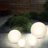 Four illuminated spherical garden lights of varying sizes are placed on a wooden deck in front of a wooden fence and green bushes. Produktbild-Vorschau