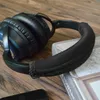 A black headphone headband cover is placed on top of a book, with headphones and a smartphone visible in the background. Produktbild-Vorschau