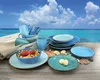 A collection of blue and turquoise ceramic dishes, cups, and bowls are arranged on a wooden table with a tropical beach and ocean in the background. Produktbild-Vorschau