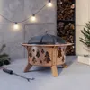 A fire pit with a Christmas tree cutout design is shown in a cozy, indoor setting with string lights and a stack of firewood. Produktbild-Vorschau