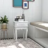 A white nightstand with a drawer sits next to a built-in bench in a room with white paneled walls and a patterned rug. Produktbild-Vorschau