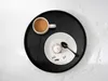 A top-down view of a black tray with a white mug of coffee and a dessert plate with a small cake and spoon. Produktbild-Vorschau