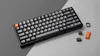 A black mechanical keyboard with gray and orange keycaps is displayed on a gray and dark gray background. Several loose keycaps are scattered around the keyboard. Produktbild-Vorschau