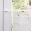 A close-up view of a white adjustable curtain rod installed on a white window frame, holding a sheer white curtain with embroidered floral patterns. The background shows a blurred view of greenery outside. Produktbild-Vorschau