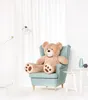 A large teddy bear sits in a light blue armchair in a room with white curtains. Anteprima dell’immagine prodotto