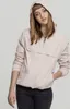 A young woman wearing a light pink hooded windbreaker and dark jeans, posing against a plain light gray background. Produktbild-Vorschau