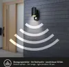 A black outdoor wall light with a motion sensor is mounted on a dark gray, horizontally-paneled wall. White curved lines emanate from the light and sensor, indicating the motion detection range. A wooden door is visible to the left. Produktbild-Vorschau