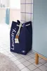 A navy blue laundry basket with white text is placed on a tiled floor next to a light blue cabinet in a bathroom setting. Náhled obrázku produktu