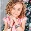 A young girl with curly blonde hair smiles at the camera, wearing a pink dress and two charm bracelets on her wrists. A decorated Christmas tree is visible in the background. Náhľad obrázku produktu
