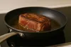 A steak is being seared in a frying pan on a stovetop. Produktbild-Vorschau