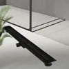 A black linear shower drain is shown on a tiled floor, with a glass shower enclosure in the background. Náhľad obrázku produktu