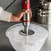 A hand holding a red and silver immersion blender over a clear plastic bowl filled with flour, with a clear plastic lid on the bowl. Produktbild-Vorschau