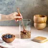 A hand is shown stirring granola in a glass jar with a wooden spoon. A bowl of granola with blueberries and a wooden lid are also visible. Produktbild-Vorschau