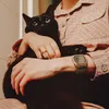 A person holding a black cat while wearing a pink striped shirt and a silver digital watch. Voir les images du produit