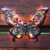 A decorative butterfly wall light with colorful jewel accents is mounted on a wooden fence. Produktbild-Vorschau