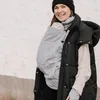 A woman wearing a black puffer vest and a black beanie is holding a baby in a gray cover. The baby is nestled inside the cover, with its head peeking out. The woman is smiling and looking at the camera. The background is a white wall. Náhled obrázku produktu
