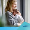 A mother holds her newborn baby in her arms, looking down at the baby with a gentle smile. The scene appears to be indoors, near a window with soft light filtering in. A blue and teal graphic overlay with wavy lines and arrows is present in the bottom righ Produktbild-Vorschau