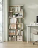 A modern, light wood bookshelf filled with books and decorative items, placed next to a window and a desk. Náhled obrázku produktu