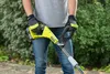 A person wearing gardening gloves and jeans is holding a yellow and black electric trimmer in an outdoor setting with greenery in the background. Podgląd zdjęcia produktu