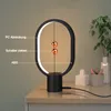 A modern black oval-shaped LED desk lamp with two magnetic spheres levitating in the center, placed on a wooden table next to decorative items and a plant. Produktbild-Vorschau