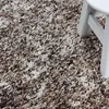 A close-up view of a textured rug with a speckled pattern of beige, brown, and white fibers. A portion of a white stool is visible in the upper right corner. Produktbild-Vorschau