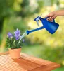 A person is watering a potted plant with a blue watering can on a wooden table. Produktbild-Vorschau