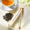 A cup of tea, loose tea leaves, and a tea water thermometer are arranged on a white surface with some greenery in the background. Produktbild-Vorschau